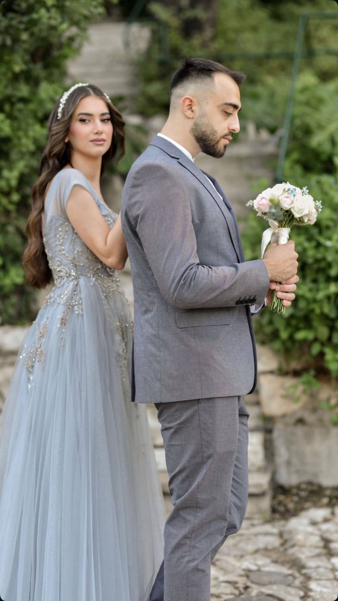 Bride in a dusty blue gown standing back to back with the groom holding a bouquet