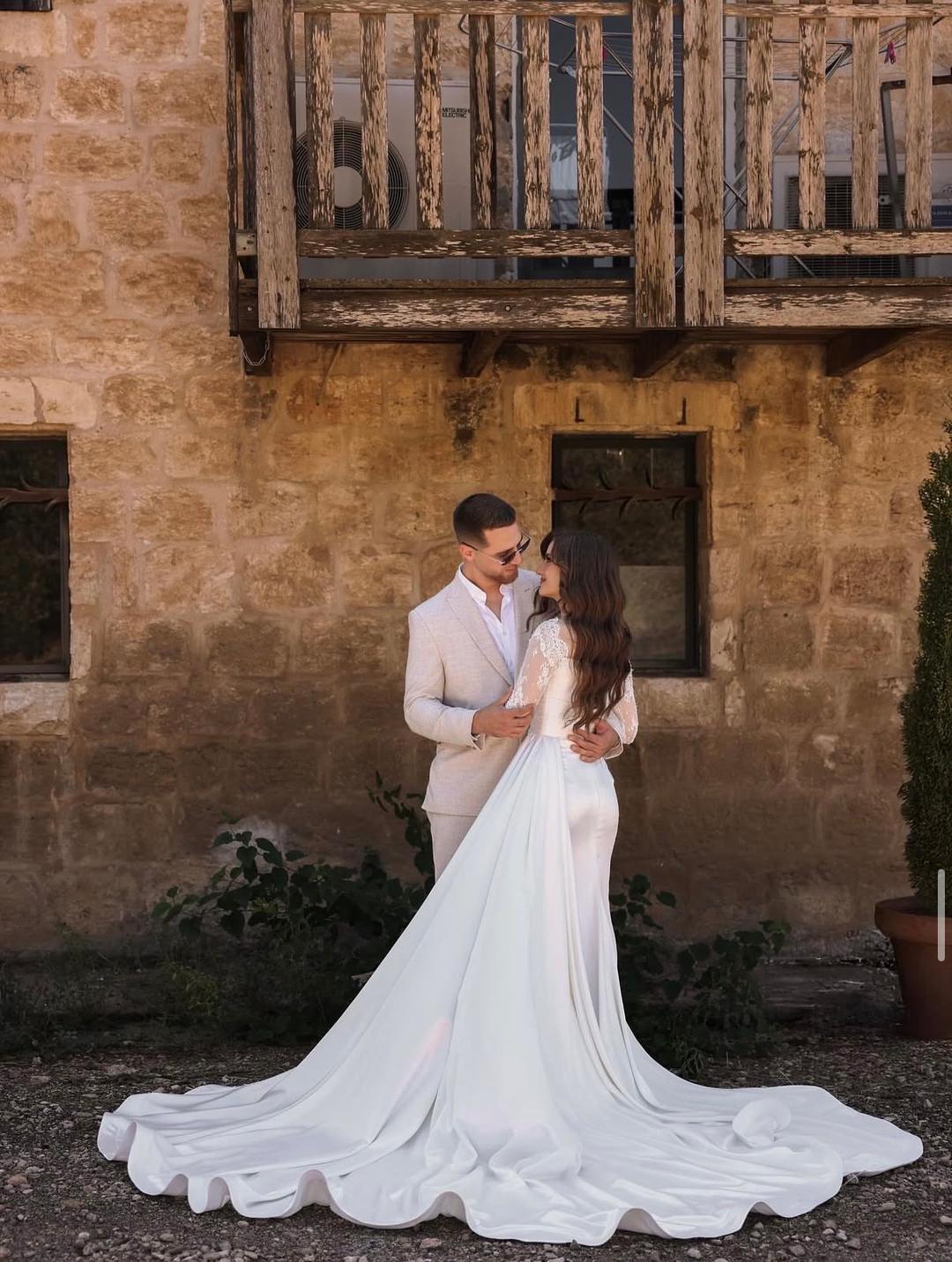 Bride and groom in cream suits embracing in front of a sunlit stone building